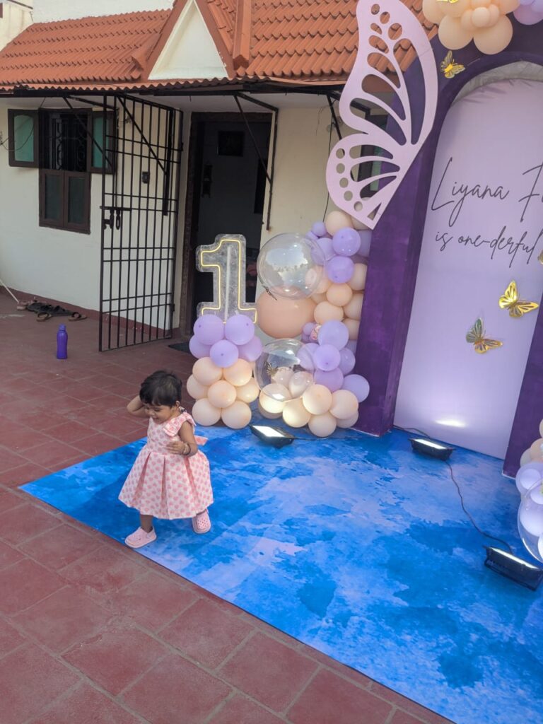 Baby girl standing near decorated terrace stage with balloons and butterfly props.