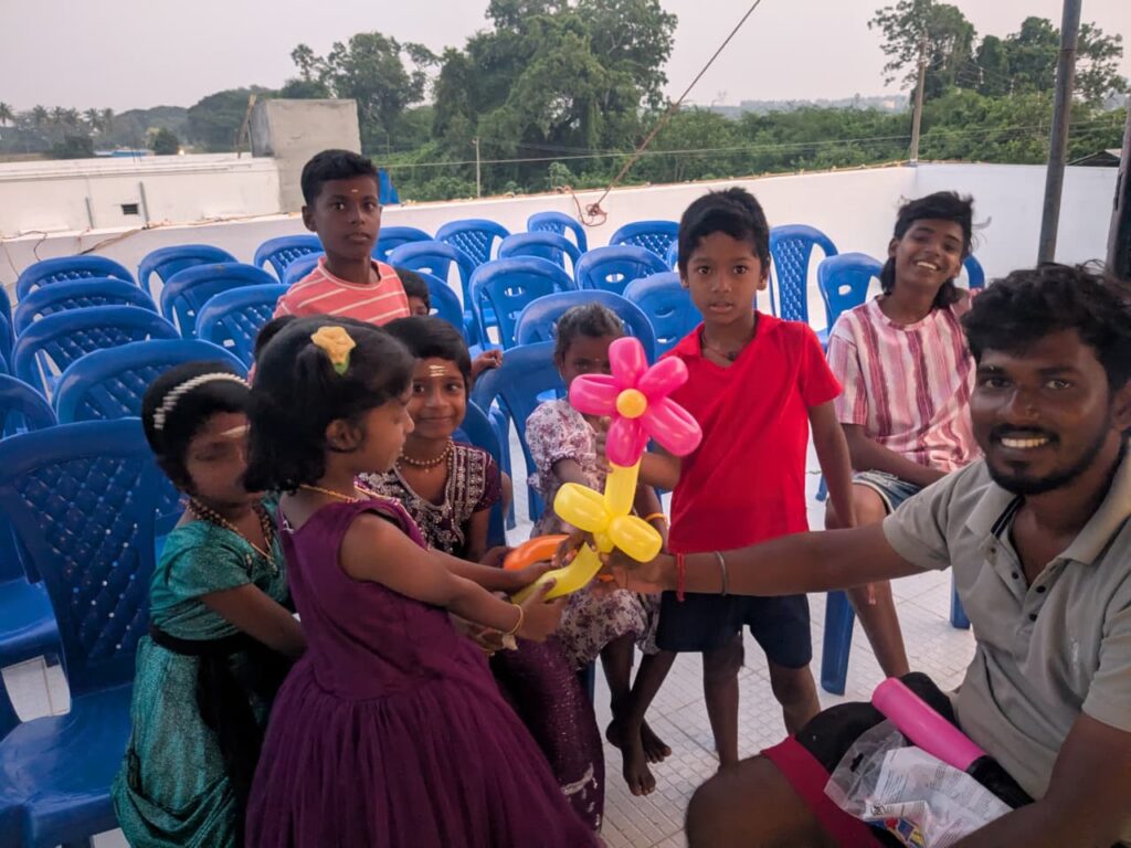 Children receiving balloon flowers and interacting with a balloon artist during Kashvik’s birthday celebration in Nolambur.
